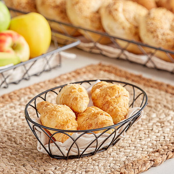 A Tablecraft round wire basket filled with croissants and fruit on a table in a bakery.