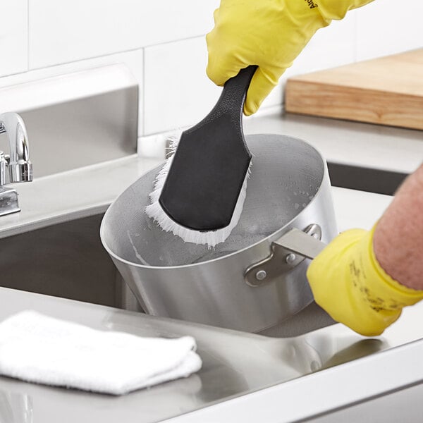 gloved hand cleaning a pan with scrub brush with black handle and white bristles