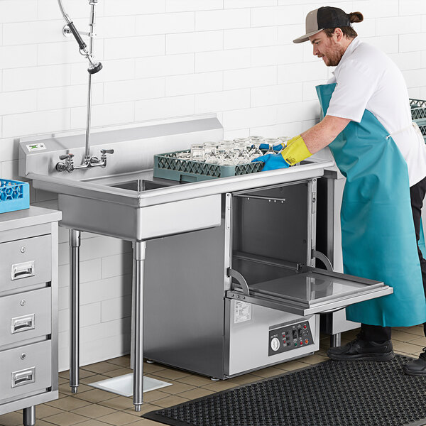 A man in a blue apron washing dishes in a sink on a stainless steel dishtable.