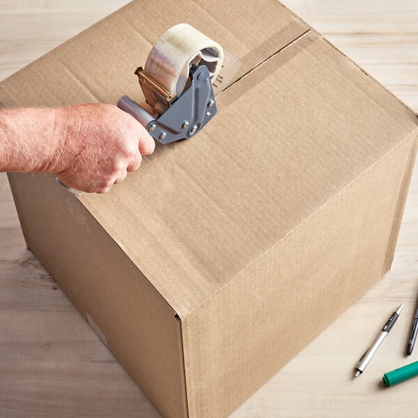 A hand using a tape dispenser to seal a Lavex Kraft corrugated shipping box.