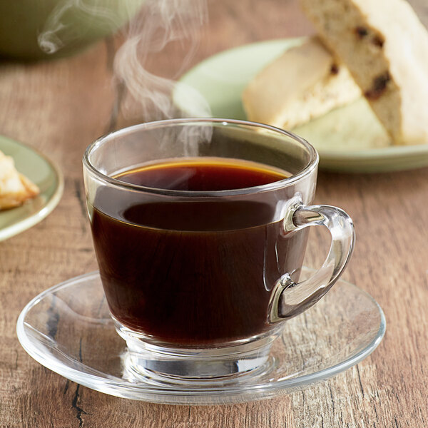 A glass cup of Ellis Mezzaroma Decaf Heavenly Hazelnut coffee on a saucer with a small piece of bread.