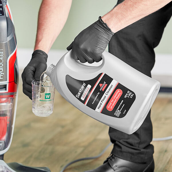 A person holding a white bottle of Sanitaire Hard Floor Cleaner pouring liquid into a beaker.