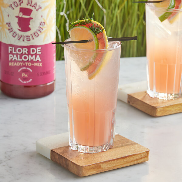 A glass of Top Hat Provisions Flor De Paloma drink with orange slices and a straw on a wooden coaster.