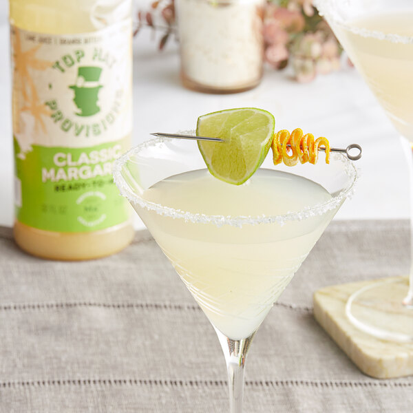 A glass of white margarita with a lime on the rim next to a bottle of Top Hat Provisions Classic Margarita Mix.