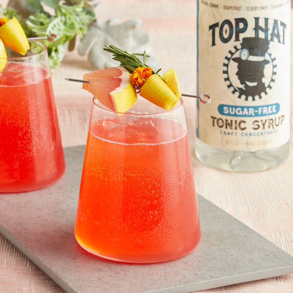 A glass of red liquid with a fruit garnish and a yellow flower next to a bottle of Top Hat Provisions Sugar-Free Elderflower and Grapefruit Tonic.