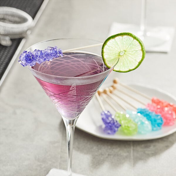 A glass of purple liquid with a lime slice on a stick next to a plate of candy.