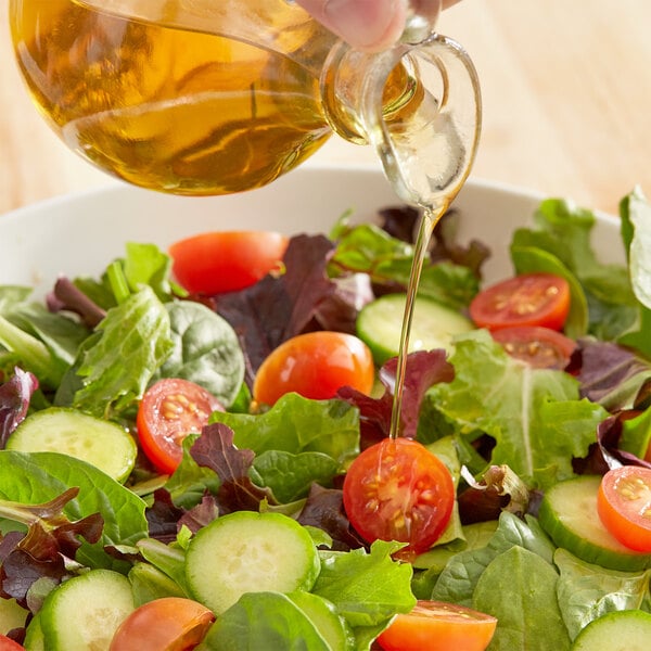 A hand pours yellow Pure Avocado Oil into a salad bowl with tomatoes, cucumbers, and lettuce.