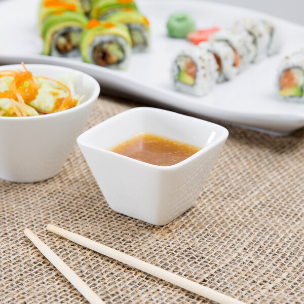 A plate of sushi with a white square porcelain sauce cup filled with brown sauce and chopsticks.