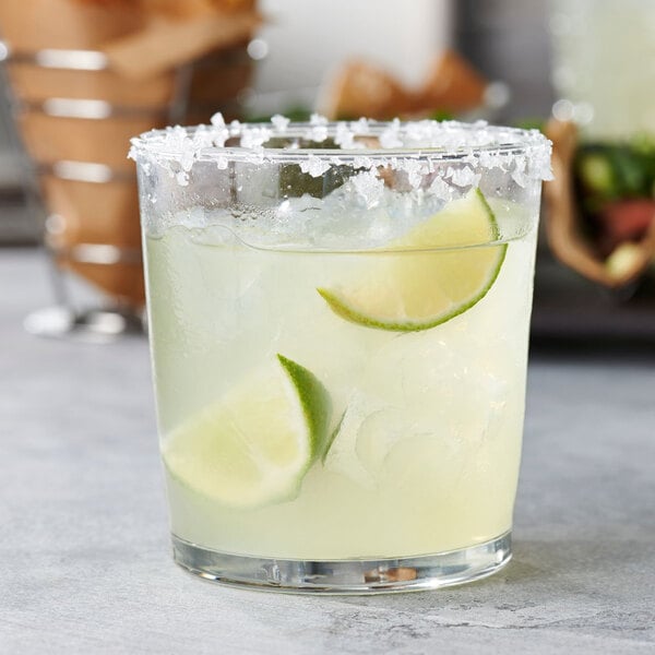 A clear glass tumbler filled with a light yellow beverage, ice, lime wedges, and a salted rim.