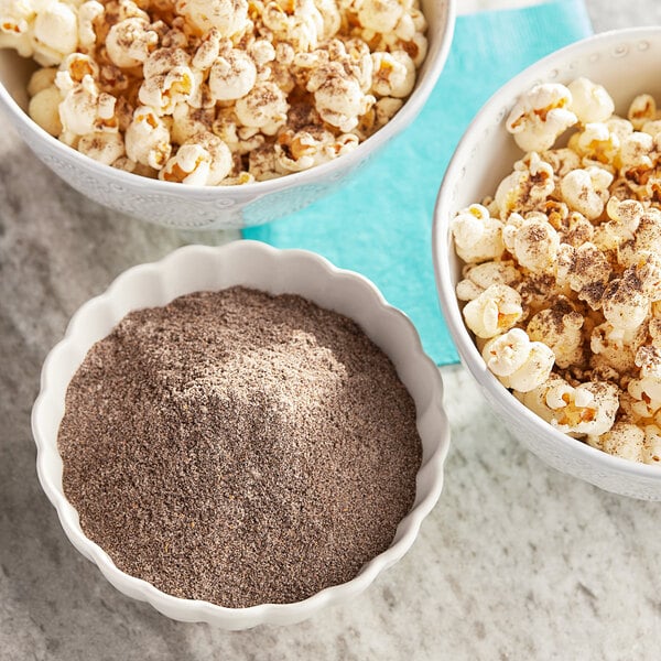 A bowl of popcorn with brown chili powder sprinkles next to a bowl of Carnival King Chili Popcorn Seasoning powder.