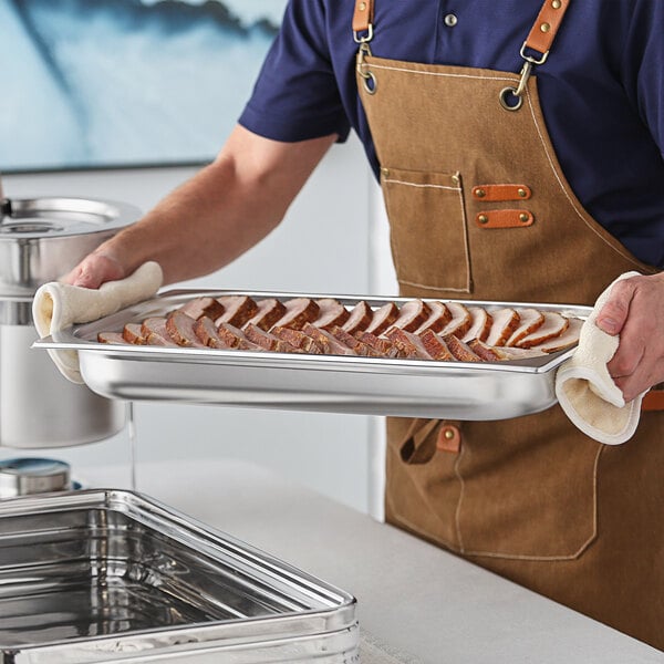 A man in an apron holding a Acopa Voyage full size chafer food pan full of food on a metal tray in a professional kitchen.