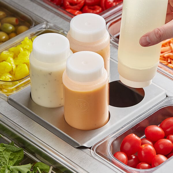 A hand using a stainless steel squeeze bottle to pour dressing into a salad container on a counter.