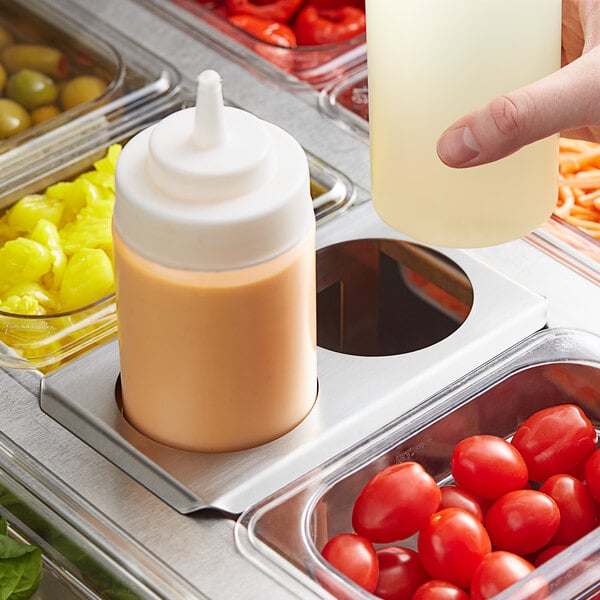 A person using a Choice stainless steel squeeze bottle holder to pour sauce into a container of food on a counter.