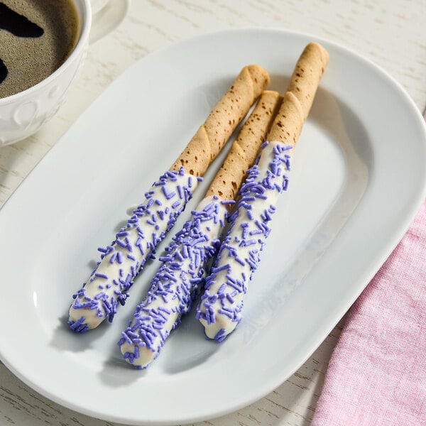 A plate with three cookie sticks partially coated in white chocolate and decorated with purple sprinkles.
