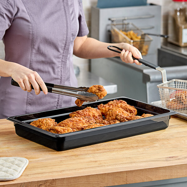 A woman using tongs to serve fried chicken from a Cambro black plastic food pan on a counter.