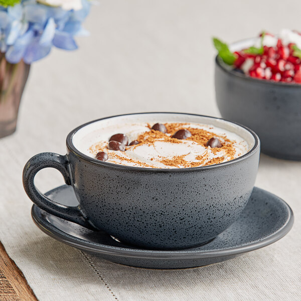 A matte midnight blue stoneware cup with a matching saucer, filled with a creamy beverage and garnished with chocolate chips and a sprinkle of cinnamon.