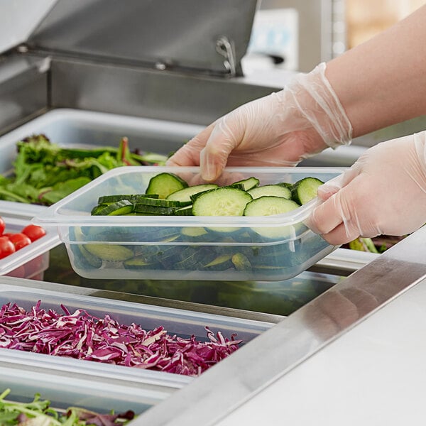 A person in gloves holding a Vigor translucent polypropylene food container filled with cucumbers.