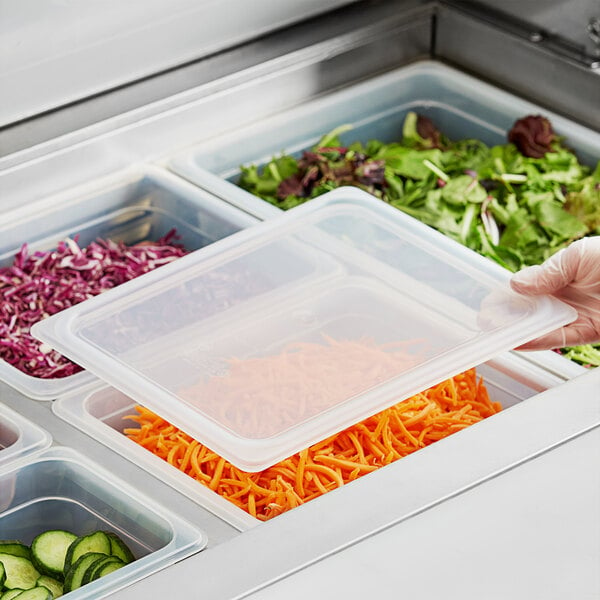 A hand in a glove securing a Vigor translucent polypropylene lid on a plastic tray of food with green leaves.