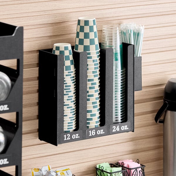 A black wall-mounted organizer with three vertical sections holding cups of different sizes labeled 12 oz., 16 oz., and 24 oz., along with a side compartment containing stirrers.
