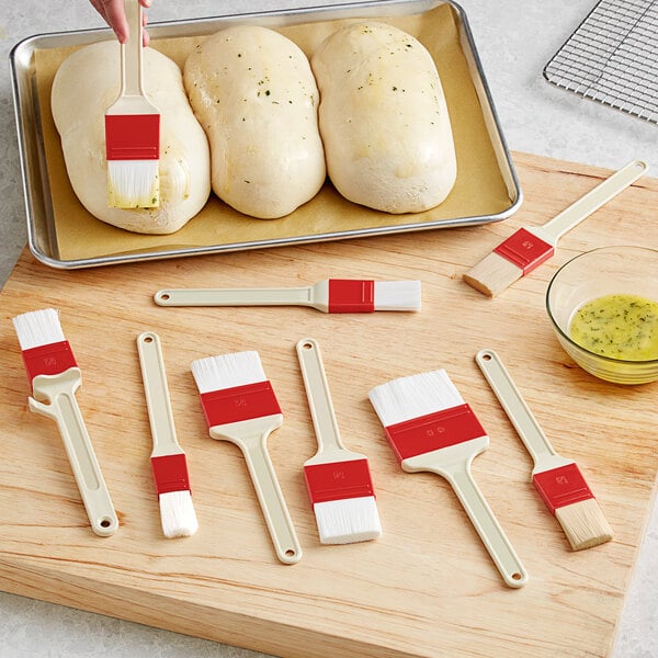 A person using a red and white Choice pastry brush to apply egg wash to bread dough on a cutting board.