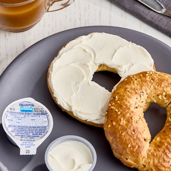 A plate with a sliced bagel, one half spread with cream cheese, accompanied by a Philadelphia Reduced Fat Cream Cheese Spread portion cup.