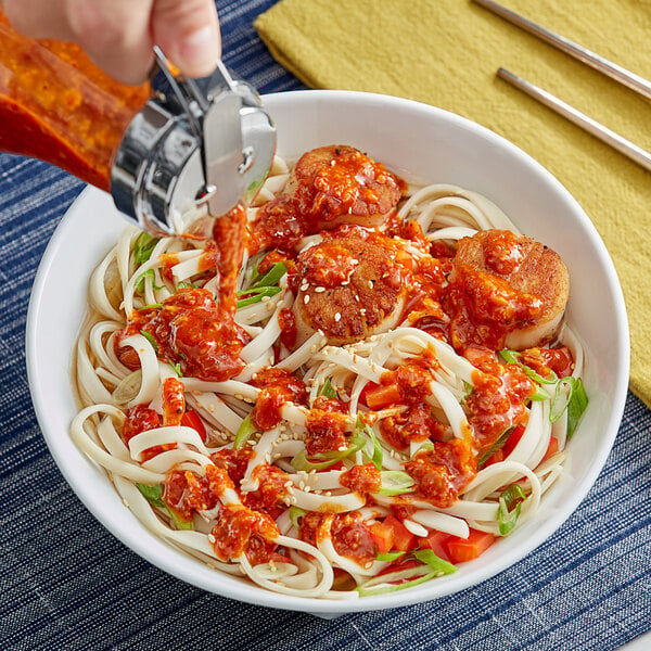 A hand pouring Ashoka Spicy Schezwan sauce into a bowl of noodles.