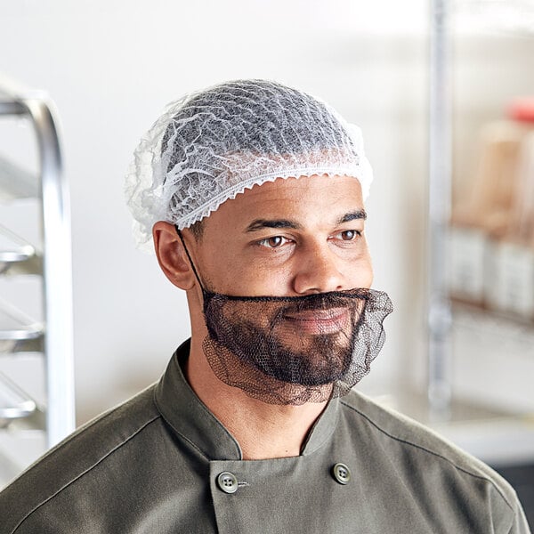 A man wearing a white disposable pleated bouffant cap on his head in a professional kitchen.