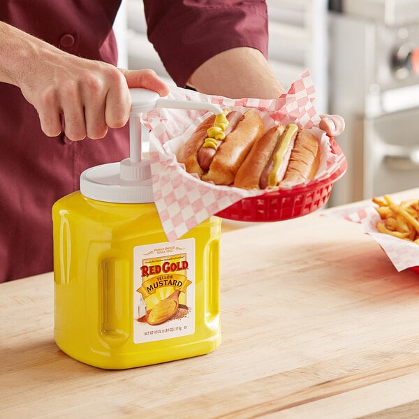 A man using a yellow plastic jug of Red Gold mustard to pump mustard onto a hot dog.