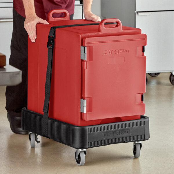 A man using a black cart to transport a large red CaterGator food pan carrier.
