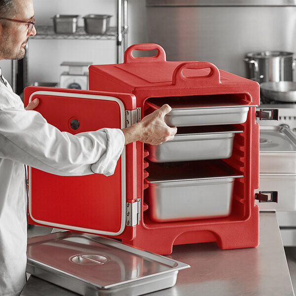 A man in a white shirt and red chef hat opening a red metal CaterGator food pan carrier.