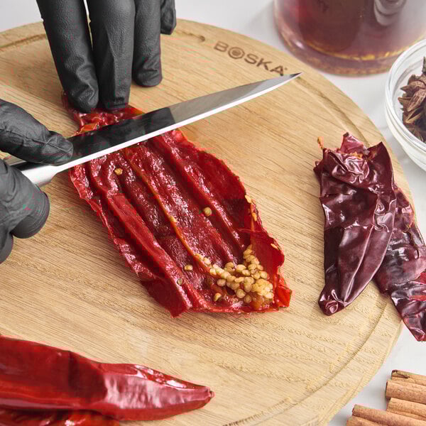 A person wearing black gloves is slicing open a dried guajillo chile pepper on a wooden cutting board.