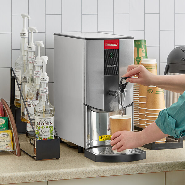 A person pouring hot water from a Grindmaster tap-operated dispenser.