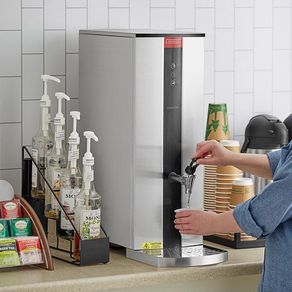 A woman using a Grindmaster tap-operated hot water dispenser to fill a cup.