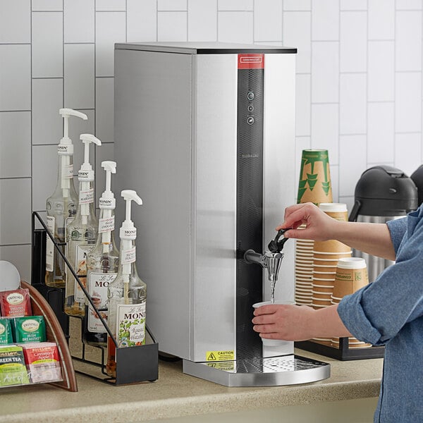 A woman using a Grindmaster tap-operated hot water dispenser to fill a coffee cup.