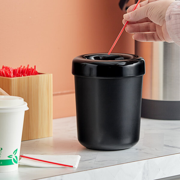 A hand holding a red straw above a black American Metalcraft round table top waste bin with a lid.