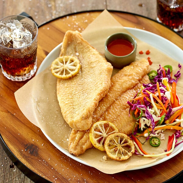 A plate of fried fish fillets served with lemon slices, coleslaw, a small cup of sauce, and a glass of iced beverage.