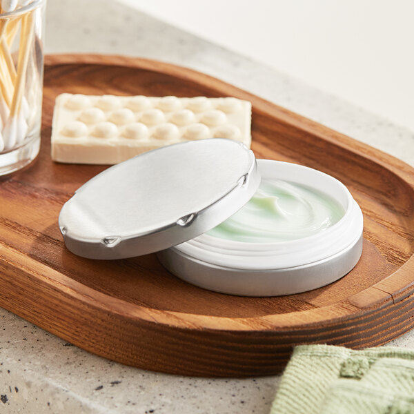 A round silver metal tin with a notched lid on a wooden tray with a jar of cream and toothpicks.