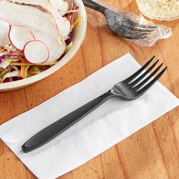 An individually wrapped Solo Impress black plastic fork on a napkin next to a bowl of salad.