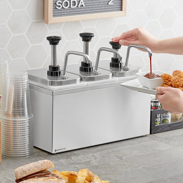 A person using a ServSense stainless steel condiment dispenser to pour sauce onto food.