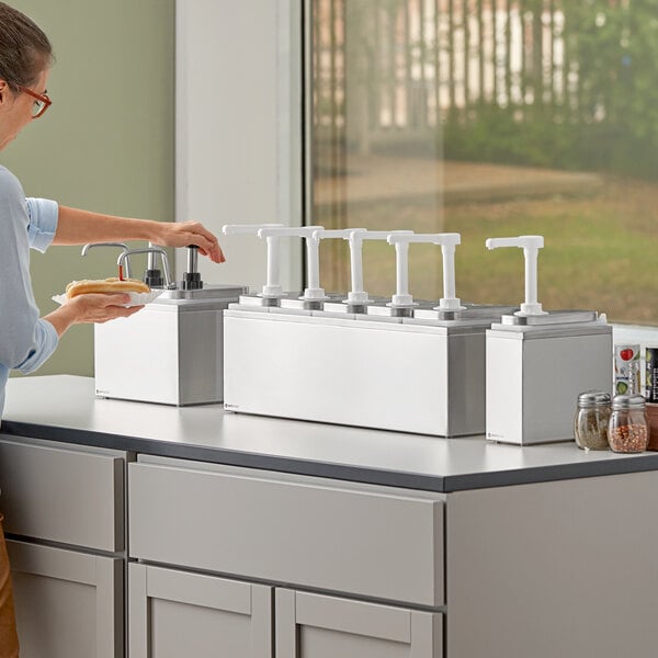 A woman using a stainless steel ServSense condiment dispenser with white pumps on a counter.