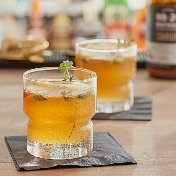 Two glasses of Apple Cardamom Shrub with a sprig of herb on top on a table.