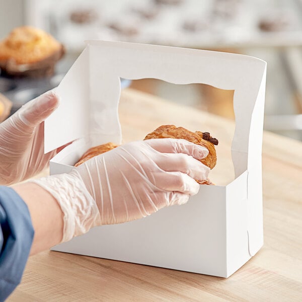 A person in pink gloves holding a Baker's Mark white window bakery box full of pastries.