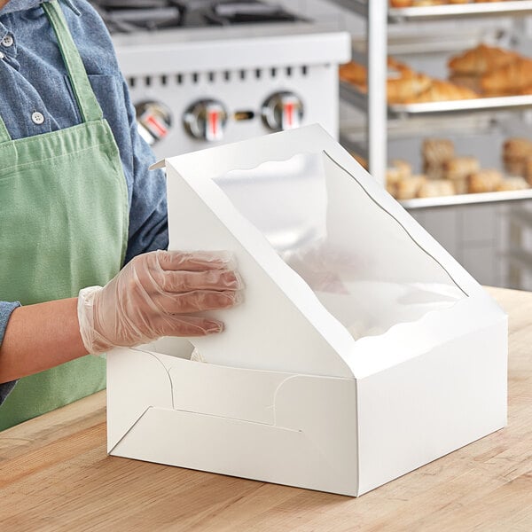 A woman wearing gloves putting a plastic bag of doughnuts into a white Baker's Mark bakery box.