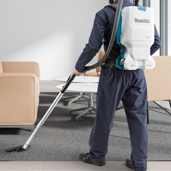 A man wearing a Makita backpack vacuum cleaner vacuuming a room.