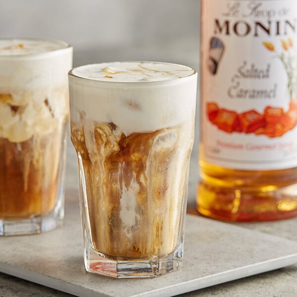 A glass of brown liquid with foam made with Monin Salted Caramel Flavoring Syrup on a table with a bottle of the syrup.