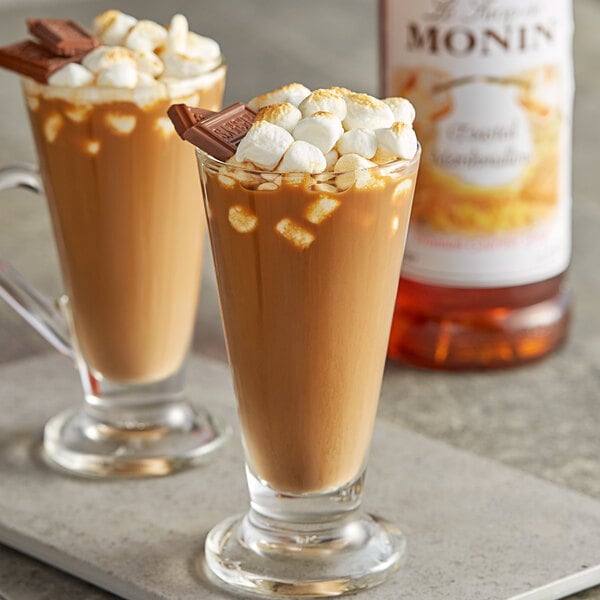 Two glasses of hot chocolate with marshmallows and a bottle of Monin Toasted Marshmallow Syrup on a table.