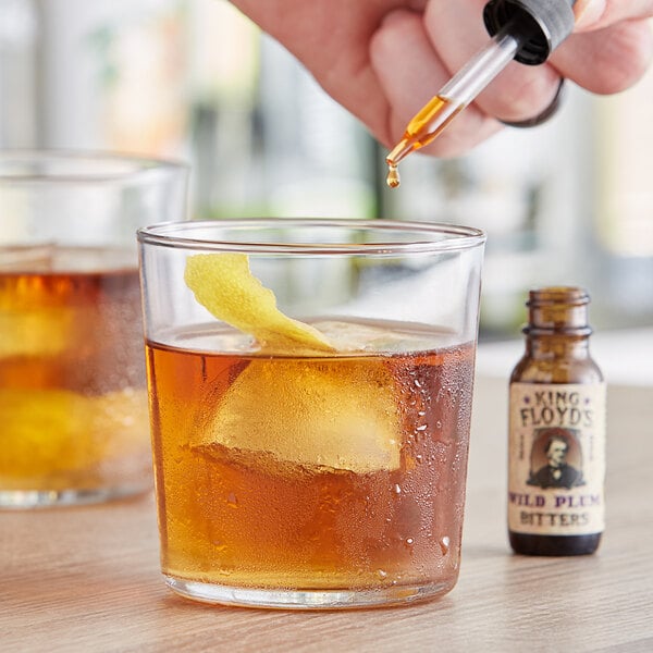 A hand using a pipette to add King Floyd's Wild Plum Bitters to a glass of brown liquid.