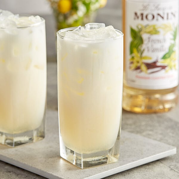 Two glasses of yellow liquid on a tray with a bottle of Monin French Vanilla Syrup.