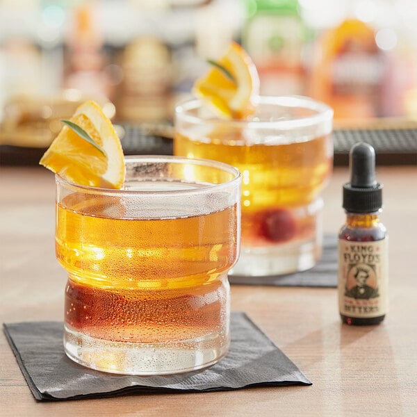 A bottle of King Floyd's Orange Bitters on a table with two glasses of liquid and orange slices.