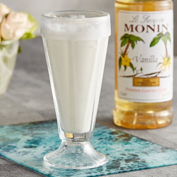 A glass of milkshake next to a bottle of Monin Premium Vanilla Flavoring Syrup.